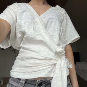 Vit omlottopp med knytband - Säljer en vit omlottopp med korta ärmar och knytband i midjan. Toppen har en v-ringad hals och ett lätt strukturerat tyg som ger en snygg look. Perfekt till jeans eller kjol!