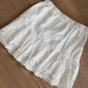 Vit kjol med broderade detaljer - Supersöt vit kjol med broderade blommor och volang längst ner. Resår i midjan för skön passform. Perfekt till sommaren och lätt att matcha med olika toppar. Storlek 158 men passar mig som har xs