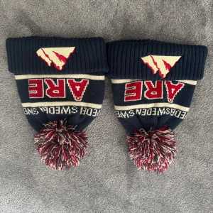 Mörkblå mössa med tofs från Åre - 200kr st. Mörkblå stickad mössa med röd, vit och blå tofs. Broderad Åre-logga framtill och texten 'Åre Sweden' runt om. Ribbad uppvikt kant och klassisk vinterdesign. Perfekt för kalla dagar. 