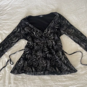 Svart omlottopp med mönster - Säljer en svart omlottopp från Benotti med långa ärmar och ett unikt grått/vitt virvelmönster. Toppen har knytband i midjan och djup v-ringning. ‼️Det står storlek 40/42 på lappen men eftersom man kan dra in den i midjan passar den även mig med S ‼️