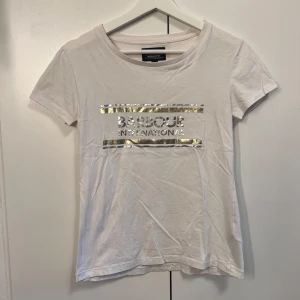 Vit t-shirt  Barbour International  - Vit t-shirt från Barbour International med korta ärmar och rund halsringning. Framsidan har ett tryck med logga och två metallicfärgade ränder i silver. Perfekt för en clean och stilren look. Använd en gång, väldigt bra skick.