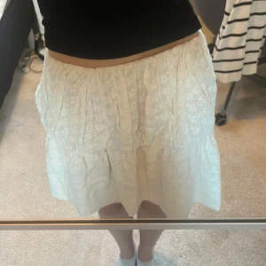 Vit volangkjol i bomull - Säljer en söt vit kjol med volanger och diskret blommigt mönster. Kjolen är kort och har en luftig passform, perfekt för varma dagar. .