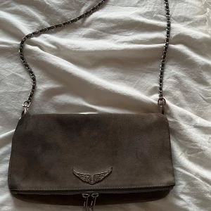 Grå axelväska i mocka från Zadig & Voltaire - Säljer en stilren grå axelväska i mocka från Zadig & Voltaire. Väskan har en kedjerem i silverfärg och dragkedja både fram och upptill. Perfekt för dig som vill ha en snygg och praktisk accessoar.