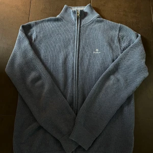 Blå stickad ziptröja från Gant - Säljer en blå stickad ziptröja från Gant med hög krage och dragkedja framtill. Tröjan har diskret broderad logga på bröstet och ribbade muddar vid ärmslut och nederkant. Perfekt för lager-på-lager under kyliga dagar.