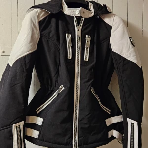 Svart och vit dun jacka från Horspist Paris - Säljer en svart och vit jacka från Horspist Paris med flera dragkedjor och snygga detaljer. Jackan har huva, markerad midja och vita partier på axlar, ärmar och rygg. Märkeslogga på ärmen och coola bältesdetaljer bak. Jackan är liten i storleken-motsvarar mindre S. Luvan är avtagbar och har stor päls om det önskas.