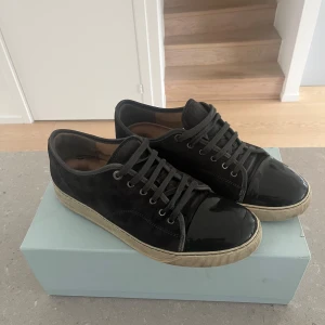Lanvin skor, svart/gråa - Säljer ett par svarta sneakers från Lanvin med snygg glansig tå. Skorna har snörning och en klassisk rund tå. Perfekta för dig som vill ha en stilren och exklusiv look. Dustbag och box ingår, finns även kvitto från care of carl