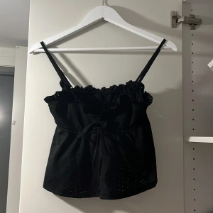 Svart linnetopp med volang och knyt - Säljer en svart linnetopp med smala axelband, volangdetalj upptill och knyt i midjan. Toppen har små broderade hål som ger en snygg touch. Perfekt för varma dagar eller lager på lager.