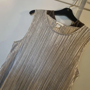 Silverfärgad plisserad klänning från H&M - Säljer en snygg ärmlös klänning från H&M i metallic silver med plisserat tyg. Klänningen har rund halsringning och rak passform, perfekt för dig som vill sticka ut med en glittrig look. Supersnygg till fest eller när du vill glänsa lite extra! Pris 100kr