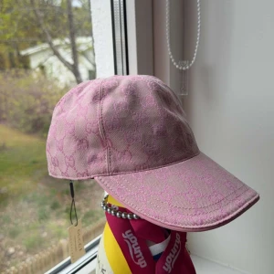 Rosa keps från Gucci med monogram - Snygg rosa keps från Gucci med klassiskt GG-monogram över hela kepsen. Justerbar rem baktill och böjd skärm. Tillverkad i bomullsmaterial och perfekt för att lyfta din stil med en lyxig touch.