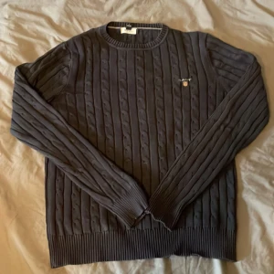 Mörkblå kabelstickad tröja från Gant - Säljer en klassisk mörkblå kabelstickad tröja från Gant med rund halsringning och broderad logga på bröstet. Tröjan har långa ärmar och ribbade muddar vid ärmslut och nederkant. Perfekt för lager-på-lager-stil.
