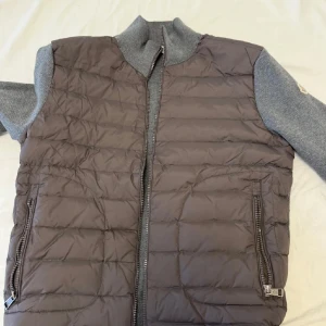 Grå cardigan från Moncler - Snygg grå cardigan från Moncler med stickade ärmar och rygg i mjuk grå ullmix. Västen har dragkedja framtill och två fickor med dragkedja. Klassisk Moncler-logga på ärmen 