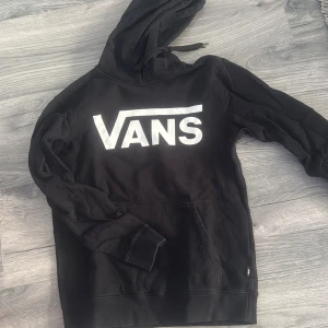 Svart hoodie från Vans - Säljer en svart hoodie från Vans med stor vit logga framtill. Tröjan har huva med dragsko och en klassisk magficka. Perfekt för dig som gillar streetwear och vill ha något bekvämt och stilrent.