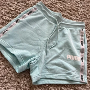 Ljusblå shorts från Puma med logoband - Snygga ljusblå shorts från Puma med vita logoband längs sidorna och dragsko i midjan. Perfekta för träning eller chill. Mjukt material och klassisk passform.
