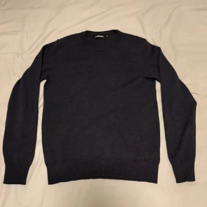 Mörkblå stickad tröja från J.Lindeberg - Säljer en stilren mörkblå stickad tröja från J.Lindeberg. Tröjan har rund halsringning och långa ärmar med ribbade muddar vid ärmslut och nederkant. Perfekt för lager-på-lager och passar till många olika outfits.