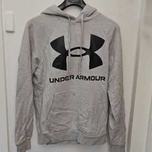 Grå hoodie från Under Armour - Grå hoodie från Under Armour med stor svart logga och text på bröstet. Tröjan har huva med dragsko och en stor magficka framtill. Perfekt för träning eller chill. Klassisk och enkel design.