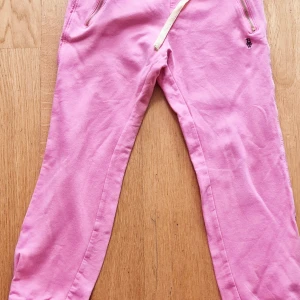 Rosa mjukisbyxor från Holly & Whyte - Säljer ett par rosa mjukisbyxor från Holly & Whyte med dragsko i midjan och guldfärgade dragkedjor vid fickorna. Byxorna har resår i midjan och raka ben. Perfekta för en avslappnad stil..  Nya aldrig använda 