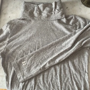 Grå polotröja DKNY - Säljer en mjuk och tunn grå polotröja från DKNY. Tröjan har lång ärm och hög polokrage, perfekt för de lite kyligare dagarna. Liten silverfärgad logga på ärmen. Stilren och enkel design.