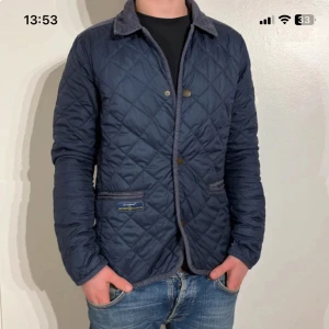Blå quiltad jacka från Barbour - Snygg blå quiltad jacka från Barbour med krage och knappar. Perfekt för höst och vår. Jackan har två fickor framtill och en stilren design som funkar till det mesta.