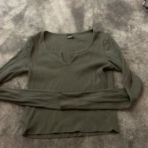 Mörkgrön ribbad långärmad topp - Säljer en mörkgrön långärmad topp med ribbad struktur och lätt v-ringad hals. Tröjan är figurnära och har en enkel, stilren design som passar till mycket. Tröjan har en liten fläck på ena armen därav det billiga priset!