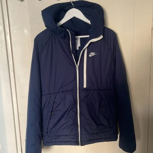 Mörkblå Therma-FIT jacka från Nike - Säljer en mörkblå Nike jacka med huva och vit dragkedja. Jackan har två fickor framtill och är tillverkad i Therma-FIT material som håller värmen. Perfekt för kyliga dagar och har en sportig look med Nike-logga på bröstet.