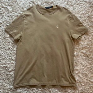 Beige t-shirt från Polo Ralph Lauren - Säljer en beige t-shirt från Polo Ralph Lauren med klassisk passform. T-shirten har korta ärmar och en liten broderad logga på bröstet. Perfekt basic-plagg med rund hals och mjukt bomullsmaterial. Pris kan diskuteras.