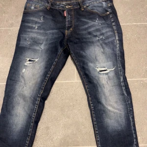 Mörkblå slitna jeans från Dsquared2 - Säljer ett par mörkblå jeans från Dsquared2 med slitningar och hål på både framsida och baksida. Perfekt skick förutom att lappen där bak har lossnat lite ( går att sy enkelt) 