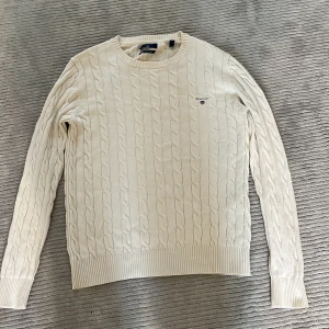 Vit kabelstickad tröja från GANT - Säljer en klassisk vit kabelstickad tröja från GANT med rund halsringning och broderad logga på bröstet. Tröjan har långa ärmar och ribbade muddar vid ärmslut och nederkant. Perfekt för en stilren och tidlös look. Storlek S men passar xs