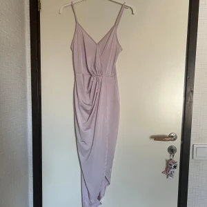 Ljuslila omlottklänning med drapering - Säljer en ljuslila klänning med smala axelband och omlott-detalj framtill. Klänningen har v-ringning och en snygg drapering längs kjolen som ger en elegant look. Perfekt för sommarens tillfällen eller balen. Jag har inte bilder på hur klänningen ser ut på men den bilden är AI och visar mycket bra på hur klänningen är. Den är glansig och inte matt som AI bilden