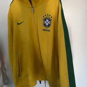 Gul Brasil jacka från Nike 2014/15 - Säljer en gul Brasil jacka från Nike med gröna detaljer på ärmarna och dragkedja framtill. Jackan har Brasil-logga och Nike-märke på bröstet samt två fickor med dragkedja. Perfekt för fotbollsälskare!