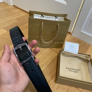 Burberry bälte - Säljer ett svart bälte från Burberry med klassiskt diskret rutigt mönster och silverfärgat spänne. Bältet har Burberry-logga på spännet och är gjort i skinn med textil på ovansidan. 3 egna gjorda hål. Spännets går att ta bort 