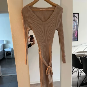 Beige ribbad långklänning med v-ringning och knytband - Säljer en beige ribbad långklänning med v-ringning och långa ärmar. Klänningen har ett knytband i midjan och en slits på sidan. Använd 1 gång och är i storlek XS 
