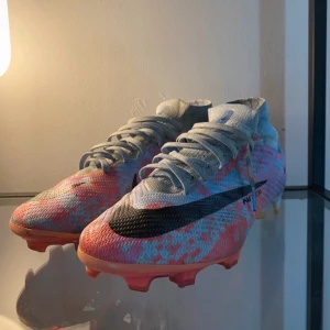 Nike Mercurial fotbollsskor med mönster - Säljer ett par Nike Mercurial fotbollsskor med grå bas och färgglada rosa och blå detaljer. Skorna har snörning, hög krage och svart Nike-logga på sidan. Perfekta för dig som vill sticka ut på planen!