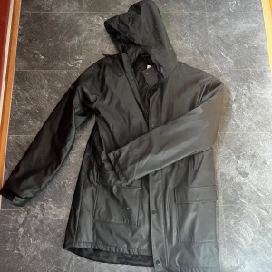 Svart regnjacka från 157 - Säljer en svart regnjacka från 157 med huva och stora fickor framtill. Jackan har knäppning framtill och är tillverkad i ett vattenavvisande material. Funkar både till killar och tjejer👋🏼