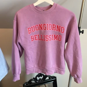 Ljuslila sweatshirt med text från Gina Tricot - Säljer en ljuslila sweatshirt från Gina Tricot med tryckt text 'BUONGIORNO BELLISSIMO' i rött och ljusrosa framtill. Tröjan har rund halsringning och långa ärmar. Perfekt för en avslappnad och trendig look.