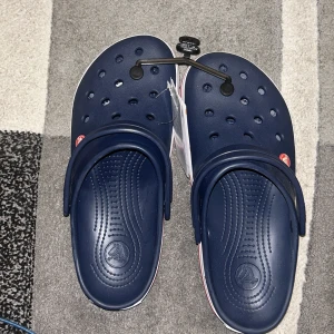 Mörkblå Crocs Crocband - OBS!! STORLEKEN ÄR MYCKET MINDRE(jag har strl 38-39 och dom passar så bra) Säljer ett par mörkblå Crocs Crocband med vit och röd rand runt sulan. Skorna har klassiska ventilationshål och justerbar hälrem. Perfekta för sommaren och riktigt bekväma att ha på sig hela dagen. Helt nya, och har aldrig använts innan!! 