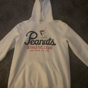 Vit Peanuts hoodie med tryck - Säljer en vit hoodie med tryck från Peanuts Athletic Club. Framsidan har en stor ficka och motiv av Snoopy med text i svart och rött. Perfekt för dig som gillar streetwear och vill ha något bekvämt och snyggt.