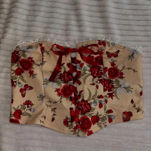 Blommig korsettopp med rosett - Supersöt korsettopp i beige med röda blommor och fjärilar, dekorerad med röd rosett framtill och spetskant upptill. Perfekt för att lyfta vilken outfit som helst!