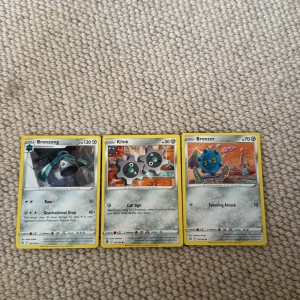 Pokémonkort: Bronzong, Klink, Bronzor - Tre Pokémonkort säljes: Bronzong med 130 HP, Klink med 50 HP och Bronzor med 70 HP. Alla kort har gul ram och klassisk Pokémon-design på baksidan. Motiven visar respektive Pokémon i färgglada miljöer. Perfekt för samlare eller Pokémon-fans!