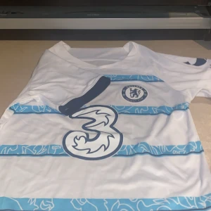 Chelsea bortatröja 2022/23 - Chelsea FC:s bortatröja från säsongen 2022/23. Vit med blåa horisontella ränder med grafiskt mönster, klubbmärke och Nike-logga på bröstet samt sponsortryck på framsidan. Kortärmad och tillverkad i lätt, ventilerande material. Storlek S passar M