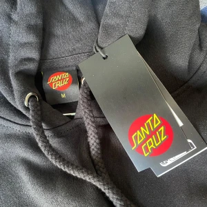 Svart hoodie från Santa Cruz - Svart hoodie från Santa Cruz med klassisk logga i rött och gult på bröstet och en stor logga på ryggen. Tröjan har huva med dragsko och en stor magficka framtill. Perfekt för dig som gillar streetwear och skate.