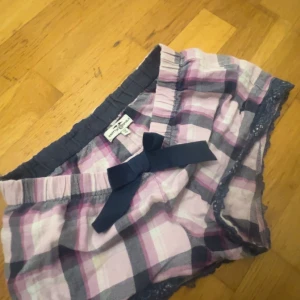 Rutiga shorts med rosett från Hampton Republic - Stolek 146/152 men passar som xs Söta rutiga shorts i rosa, grått och vitt från Hampton Republic. De har en bred resår i midjan, en stor mörkblå rosett framtill och spetsdetaljer längs bensluten. Perfekta för varma dagar eller som mysplagg hemma.