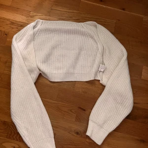 Vit stickad bolerotopp - Säljer en vit stickad bolerotopp med lång ärm. Tröjan har en kort, croppad passform och är perfekt att slänga över en topp eller klänning. Stickad struktur och breda muddar vid ärmslut.