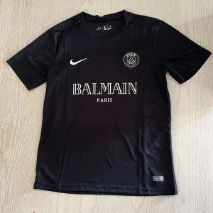 Svart PSG x Balmain fotbollströja helt ny med taggar - Snygg svart fotbollströja från Nike med Paris Saint-Germain och Balmain Paris-logga på bröstet. Tröjan har korta ärmar och är tillverkad i ett lätt och sportigt material. Perfekt för dig som gillar stilrena matchtröjor med exklusiv känsla.
