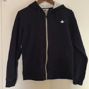 Mörkblå hoodie från Macchia J. - Mörkblå hoodie med vit dragkedja och vit stjärnlogga på bröstet. Tröjan har huva med vit insida och två fickor framtill. Säljer pga fel storlek. Använd två gånger