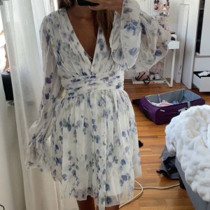 Vit blommig omlottklänning med v-ringning - Intressekoll på den populära nakd klänningen som är helt slutsåld. Romantisk vit kortklänning med blått blommönster, djup v-ringning och omlottliv. Klänningen har långa, vida ärmar med volangdetaljer och markerad midja. Perfekt för sommarens tillfällen.