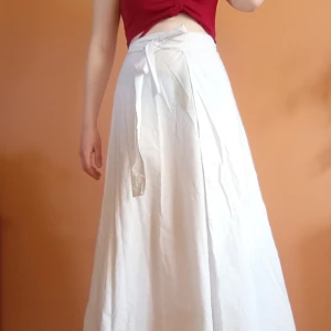 Vit omlottkjol från Tanzania  - Vit omlottkjol med knytband i midjan. Kjolen är ett långt tyg som man sedan med hjälp av banden snurrar runt midjan och knyter i en rosett. A-linjeform och tyget har tunna grå ränder. Finns en liten fläck - se bild.  Passar alla storlekar 🤍