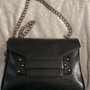 Svart axelväska med kedja från Tiamo - Säljer en svart axelväska från Tiamo med snygg kedjerem och strukturmönster som påminner om ormskinn. Väskan har ett lock med dekorativa nitar och en innerficka med dragkedja. Perfekt för att lyfta vilken outfit som helst.