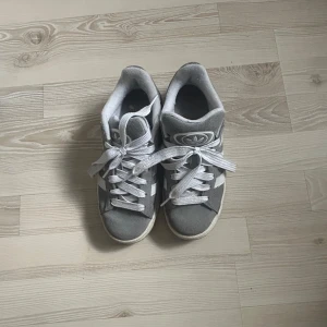 grå adidas campus skor - Säljer ett par grå Adidas Campus sneakers med klassiska vita ränder och vit sula. Skorna har snörning och är i mocka med Adidas-logga på plösen och hälen. Perfekta för en stilren och avslappnad look.