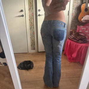 Lågmidjade blå jeans med broderade detaljer - Säljer ett par lågmidjade blå jeans med bootcut passform. Jeansen har broderade detaljer på bakfickorna, ena med strass och andra med rosa broderi. Passar mig som är 165cm 77innerben å 78 midja
