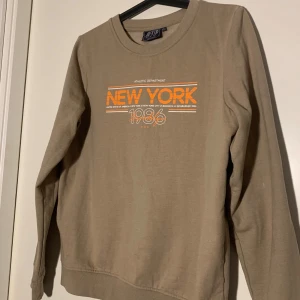 Beige sweatshirt - Säljer en beige sweatshirt från OTP med orange och vitt tryck på bröstet där det står 'NEW YORK 1986'. Tröjan har rund halsringning och långa ärmar. Perfekt för en avslappnad stil.